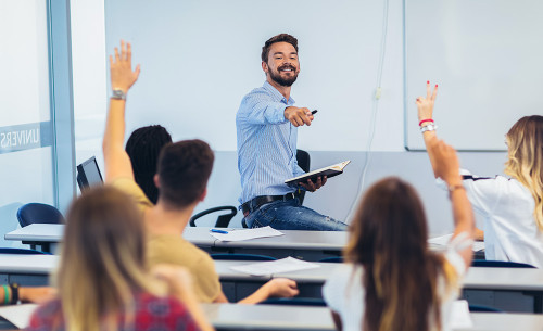 Ein Lehrer unterrichtet eine Klasse, wo Schülerinnen und Schüler gerade aufzeigen.
